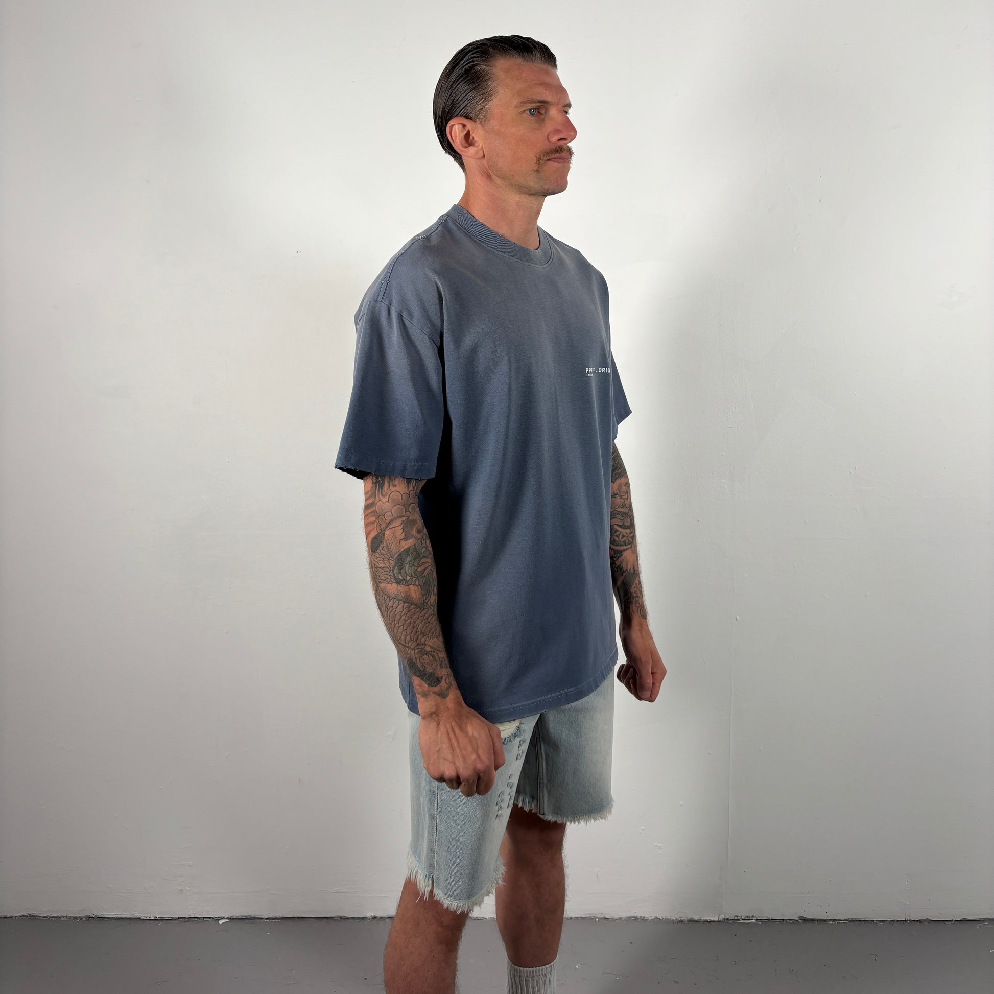 Man wearing a blue t-shirt and light gray shorts standing against a plain white wall.
